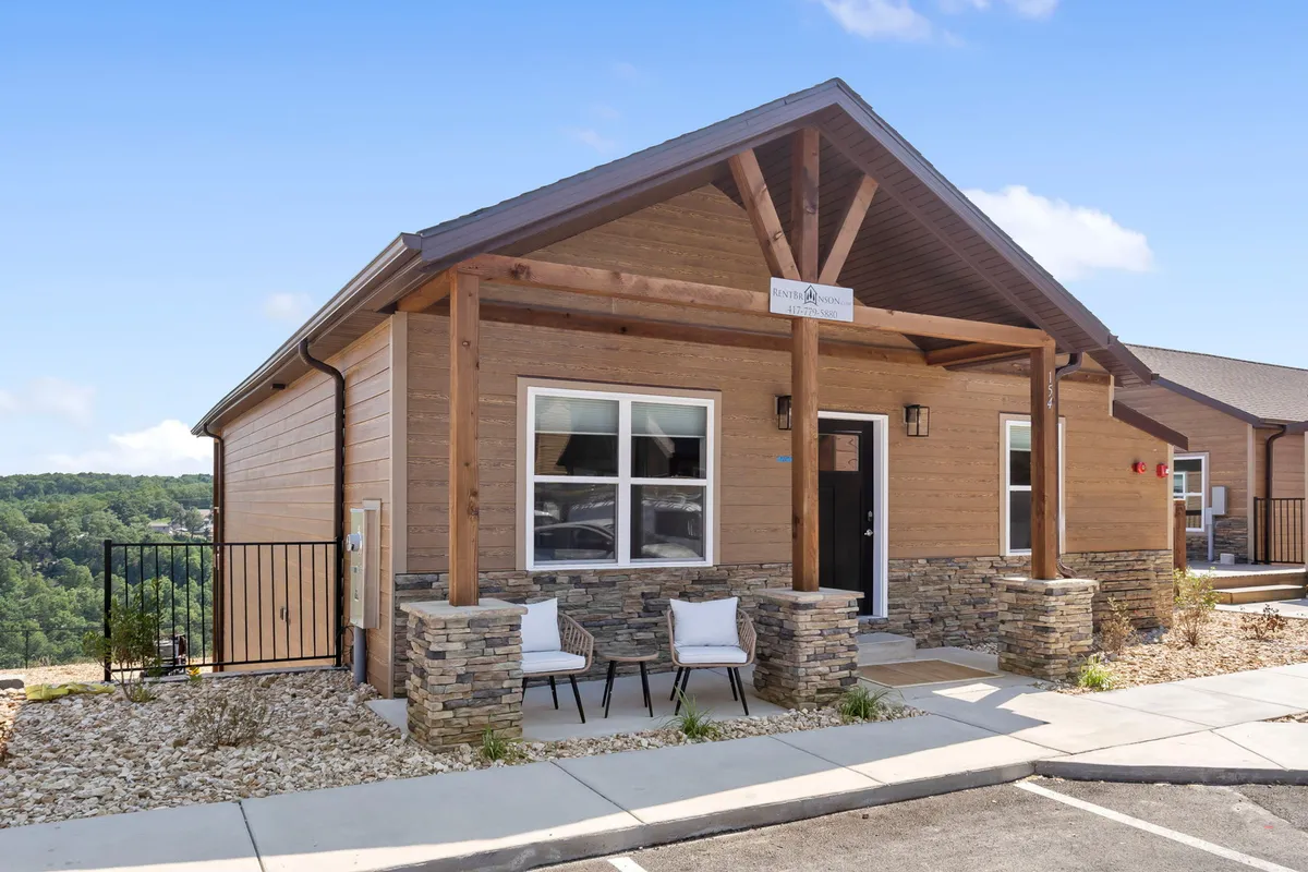 The image presents the exterior of a welcoming home, featuring wooden beams, stone accents, and inviting front porch seating. The scene conveys a sense of calm and hospitality, with space for relaxing or socializing while surrounded by nature.