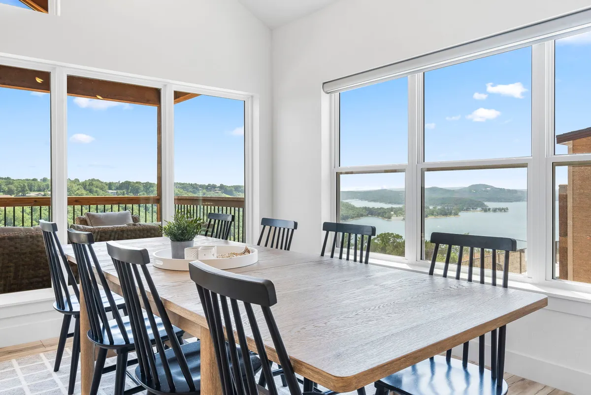 A large dining table is positioned by panoramic windows with stunning lake and green landscape views. The scene is warm and inviting, suggesting a space for family gatherings, casual conversation, and memorable holiday celebrations.