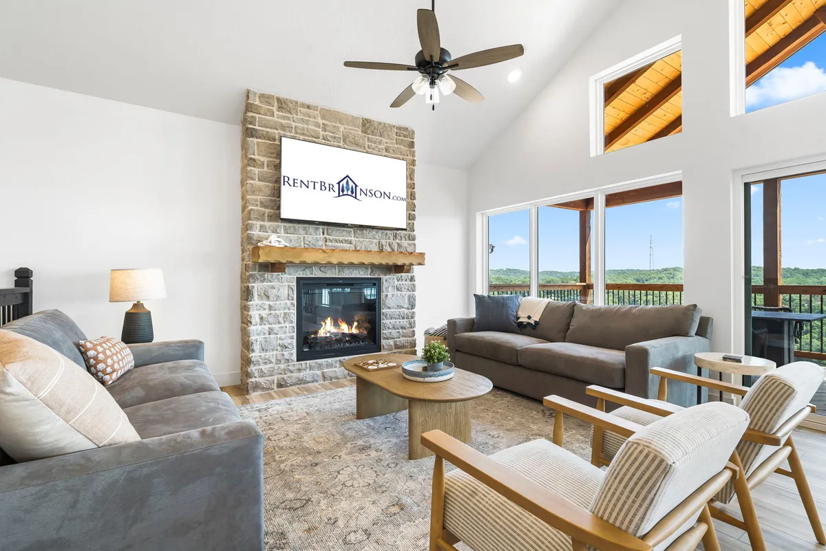 The living room features a plush sofa and cozy seating arranged around a grand stone fireplace, with large windows that frame peaceful lake views. The soft lighting and thoughtful layout create a warm, inviting atmosphere perfect for relaxing or socializing.