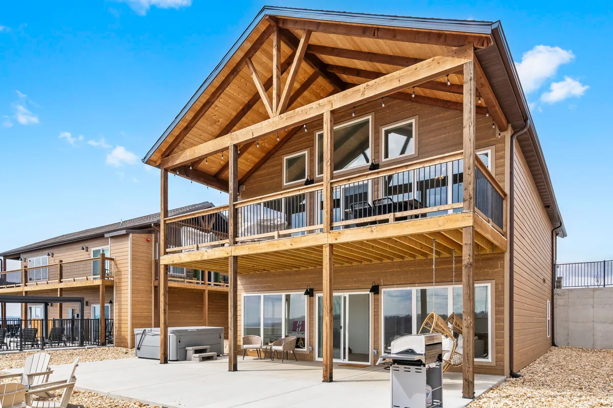 This image showcases a large deck with wooden beams, glass doors leading inside, and comfortable outdoor furniture arranged to face a stunning sunset view over the lake. The deck is spacious and inviting, suggesting ample room for gatherings or quiet relaxation.