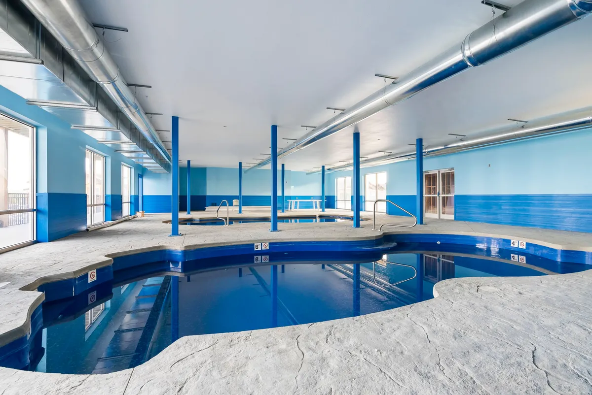 A spacious indoor pool area with bright blue walls, large windows allowing in natural light, and exposed piping for a modern touch. The inviting pool offers a comfortable place to swim and socialize regardless of the weather outside.
