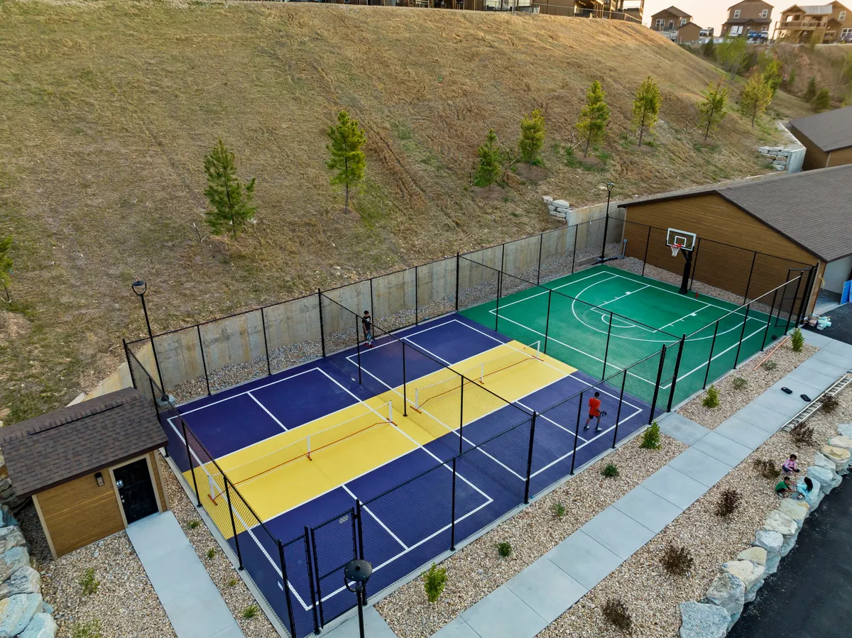 A lively exterior scene featuring multi-sport courts with guests playing basketball and pickleball, as well as a play area for children. The surrounding landscaped grounds and groups socializing create an energetic, communal atmosphere.