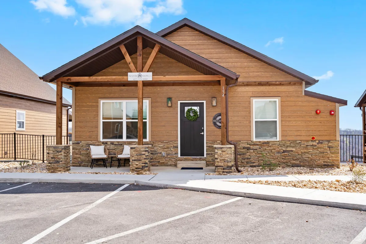 This exterior shot highlights a wooden porch with stone accents, a wreath on the front door, and comfortable outdoor seating. The setting is cozy and inviting, subtly decorated for a sense of hospitality and safety.