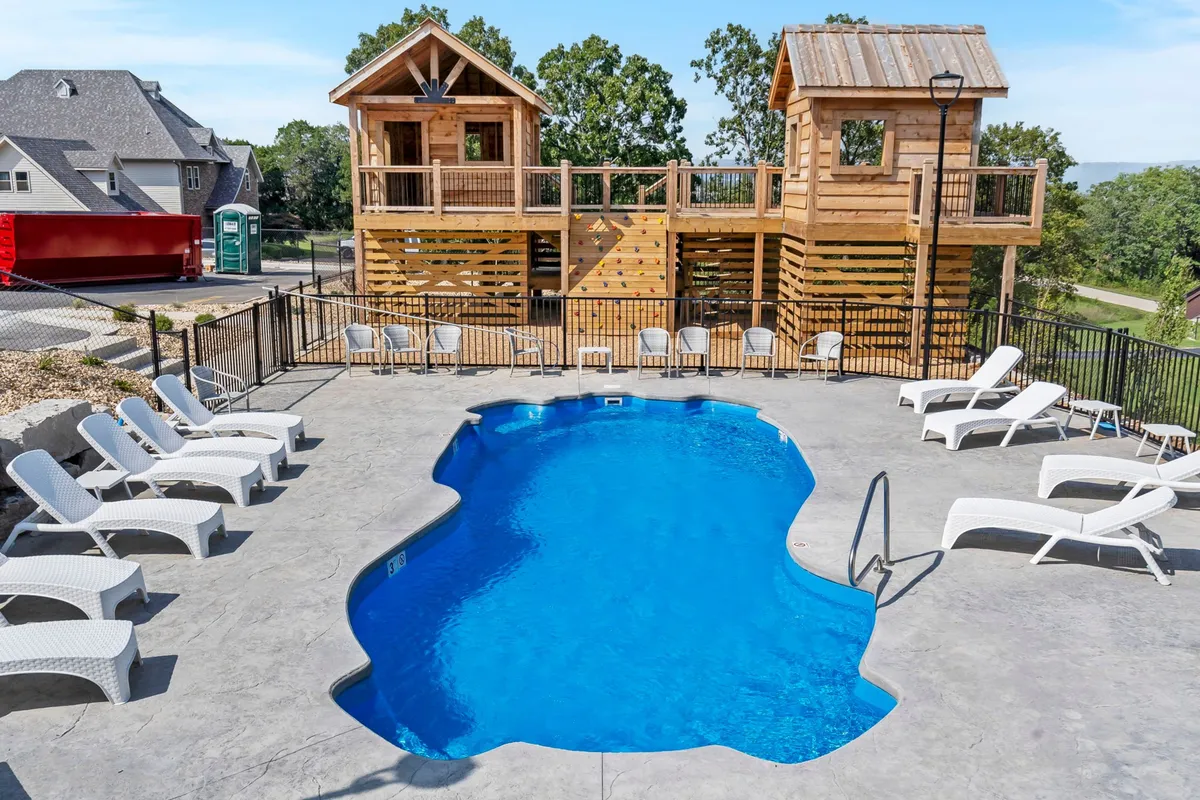 A lively outdoor scene featuring a swimming pool, wooden play structures for children, and lounge chairs. The pool area is framed by greenery and open sky, with families and friends able to gather, splash, or relax. The play structures provide a playful space for kids, while adults can unwind nearby.