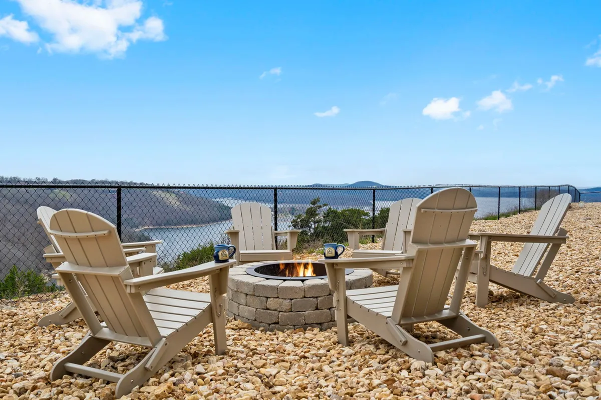 This image displays a cozy fire pit surrounded by Adirondack chairs, with a clear view of the lake beyond. The arrangement suggests intimate gatherings, stargazing, or storytelling by the fire as day turns to night.