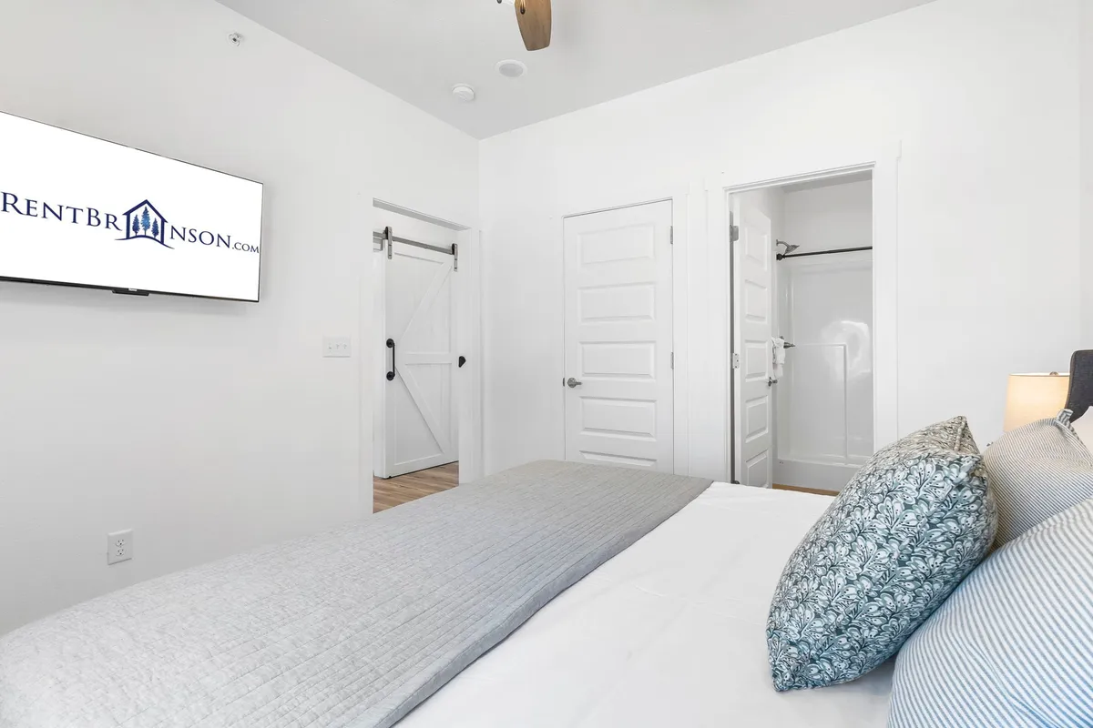 A serene bedroom with a large, comfortable bed adorned with decorative pillows, a wall-mounted television, a sliding barn door, and a closet. The room is bathed in natural light, featuring soothing colors and modern touches for a peaceful sanctuary.